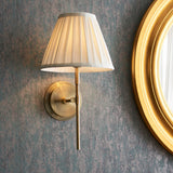 Endon Chartwell wall light with box-pleated cream cotton tapered shade on an antique brass bracket, mounted beside a gold circular mirror on a grey wall.