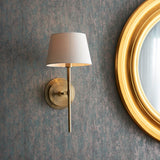 Endon Chartwell wall light with tapered ivory fabric shade on a slim antique brass bracket, mounted beside a round gold-framed mirror on a textured wall.