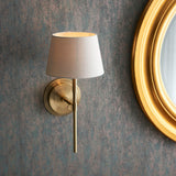 Endon Chartwell wall light with tapered ivory fabric shade and slim antique brass bracket, mounted on a textured grey wall beside a round gold mirror.
