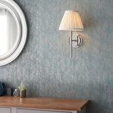 Endon Chartwell wall light in nickel with pleated cream tapered shade, mounted on a textured blue-grey wall beside a round white mirror and wooden console.