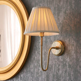 Endon Cliffdon wall light in antique brass with cream box-pleat tapered shade, mounted beside a round gold-framed mirror on a textured grey wall.