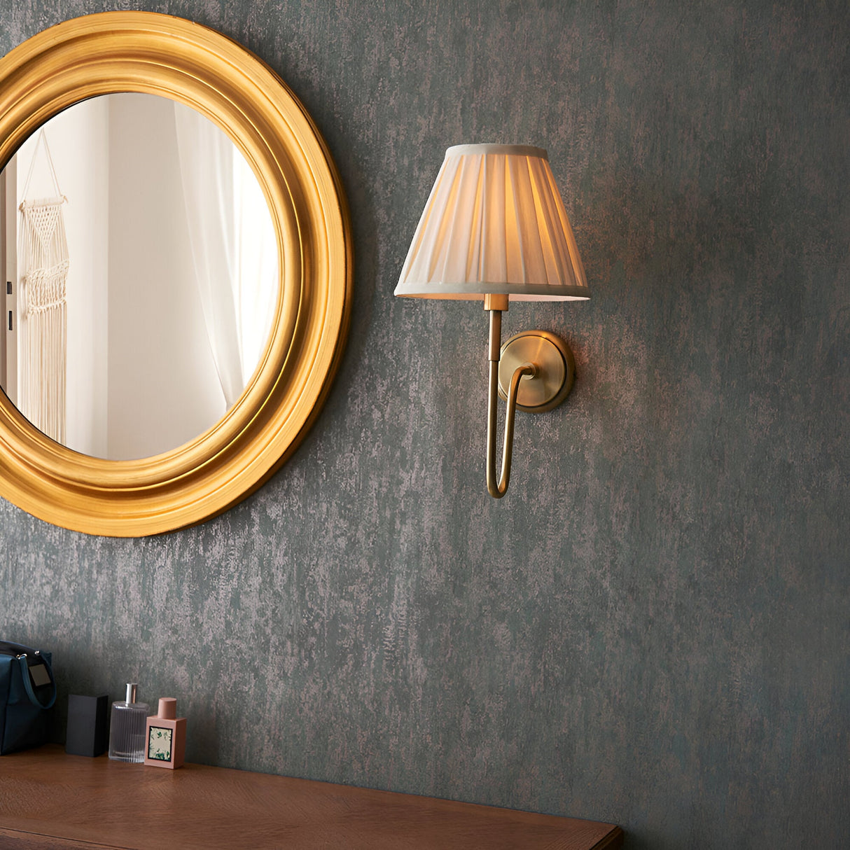 Endon Cliffdon wall light in antique brass with a cream box-pleat tapered shade, mounted beside a round gold-framed mirror on a dark textured wall.