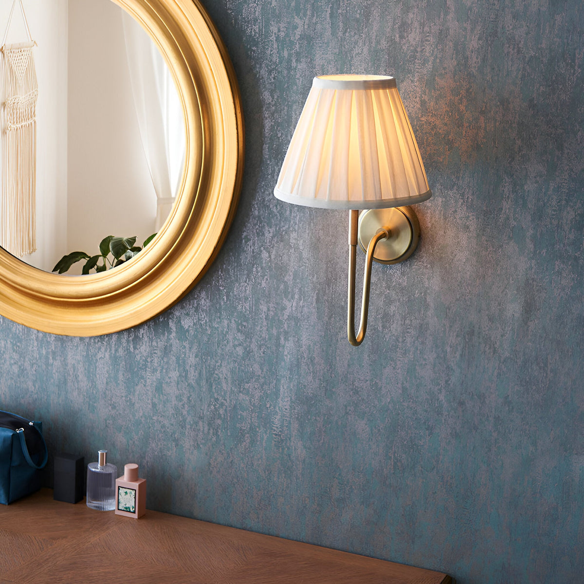 Endon Cliffdon antique brass wall light with cream box-pleat tapered shade, mounted on a textured blue wall beside a round gold-framed mirror.