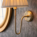 Close-up of the Endon Cliffdon antique brass wall light showing the curved arm, round backplate and pleated cream shade beside a gold-framed mirror.