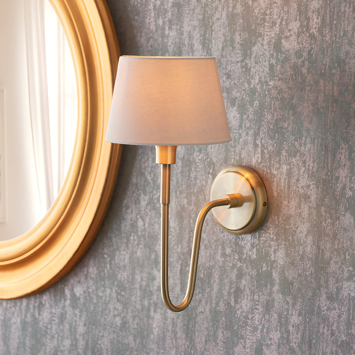 Endon Cliffdon antique brass wall light with ivory tapered fabric shade, mounted beside a round gold-framed mirror on a textured grey wall.