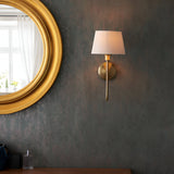 Endon Cliffdon wall light in antique brass with tapered ivory fabric shade, illuminated on a dark textured wall beside a round gold-framed mirror.