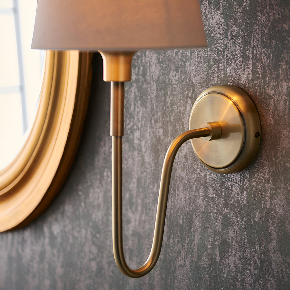 Close-up of the Endon Cliffdon wall light in antique brass with ivory tapered shade, mounted on a textured grey wall beside a gold-framed mirror.