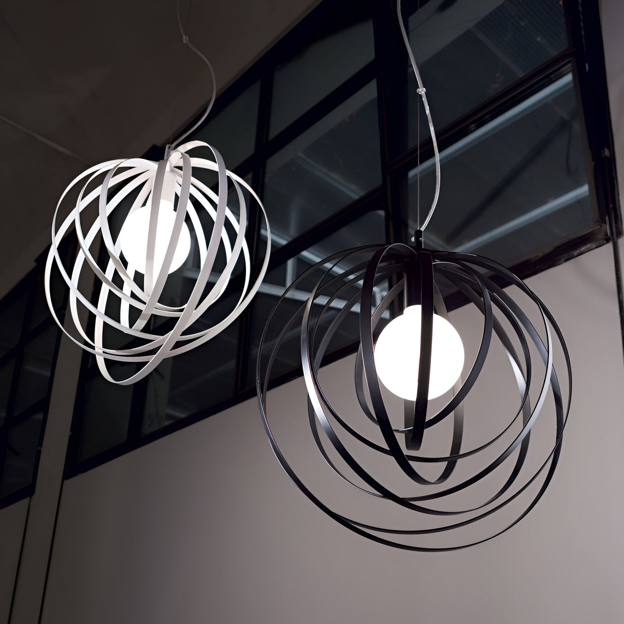 A modern interior showcases two stunning pendant lights, each with abstract design elements. The Abstract 1 Light Pendant in white features ribbon-like loops, while its counterpart boasts black loops. Both dimmable lights hang gracefully from the ceiling against a backdrop of large windows.