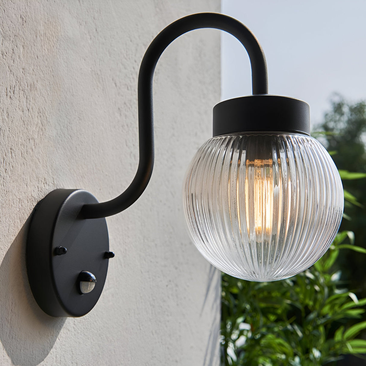 Endon Brackgrove outdoor wall light in black with arched arm, ribbed clear glass globe shade and PIR sensor, mounted on a white exterior wall.