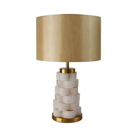 The Solmira 1 Light Stacked Alabaster Table Lamp in brass and soft white features a translucent stacked alabaster base, gold accents, and a wide cylindrical beige lampshade, displayed against a white background.