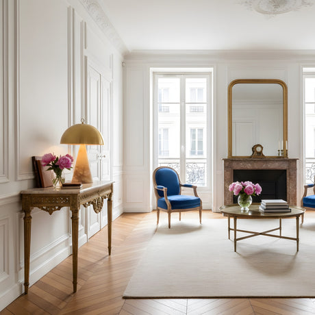 Bright, elegant living room with white walls, tall windows, a gold-framed mirror above the fireplace, blue armchairs, round coffee table with books, and a console table featuring the Viren 2 Light Polished Cap Alabaster Table Lamp in brushed bronze & soft white.