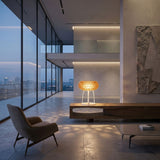 A modern, minimalist living space with floor-to-ceiling windows, a sculpted armchair, a low coffee table, and the Briller LED Table Lamp - Gold glowing on a sleek wooden console, overlooking a city skyline at dusk.