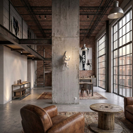 Industrial-style loft with large grid windows, exposed brick, a concrete pillar, leather armchairs, round wooden coffee table, and mezzanine with spiral staircase. The Monkey Wall Lamp - White adds sculptural character as natural light fills the spacious area.