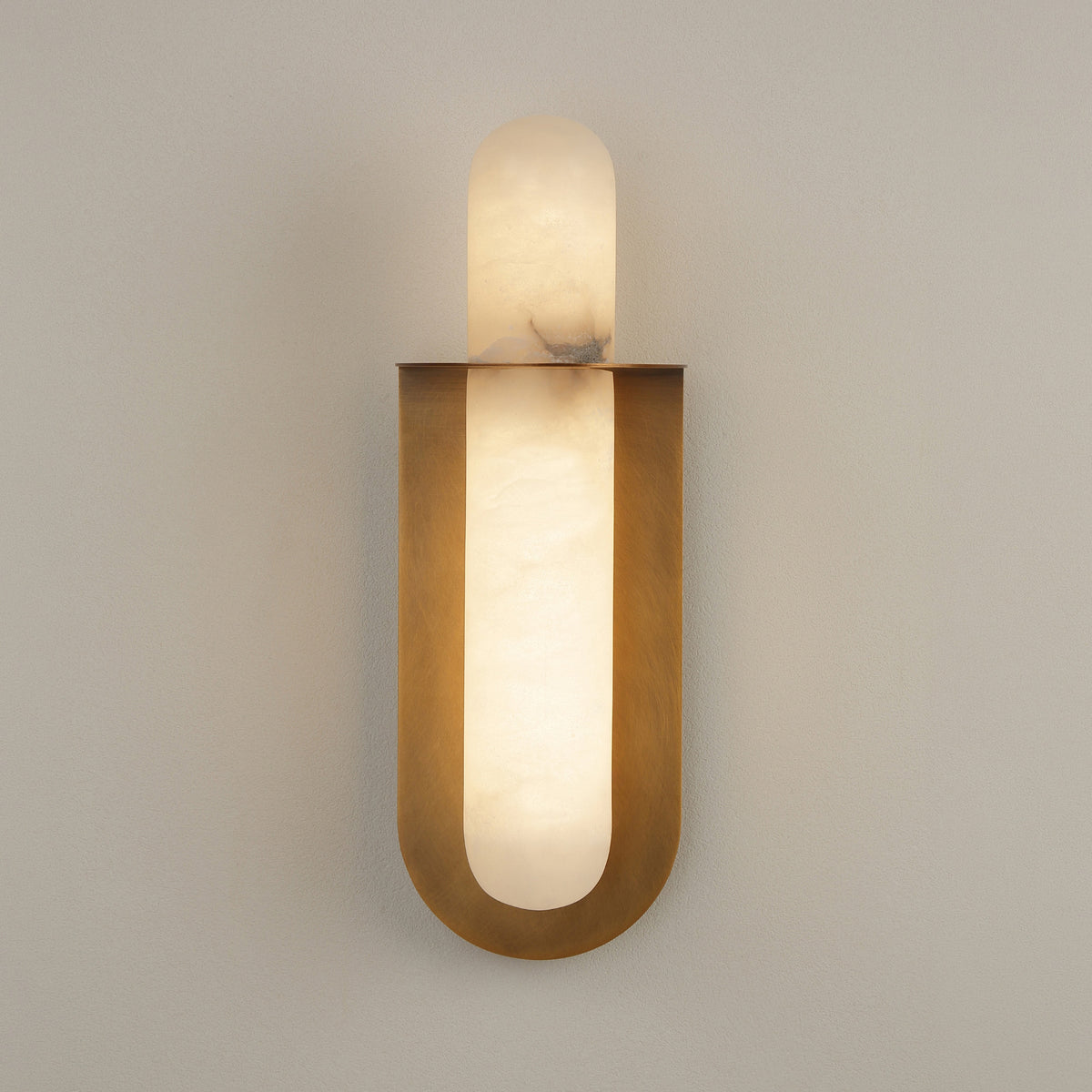 Luneth LED Small Alabaster Wall Light in Brushed Brass, with a cylindrical alabaster column set in a curved brass cradle, mounted on a plain grey wall.