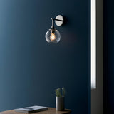 Endon Georgiana wall light in nickel with a twisted spiral clear glass globe shade, mounted on a dark blue wall above a side table with a potted plant.