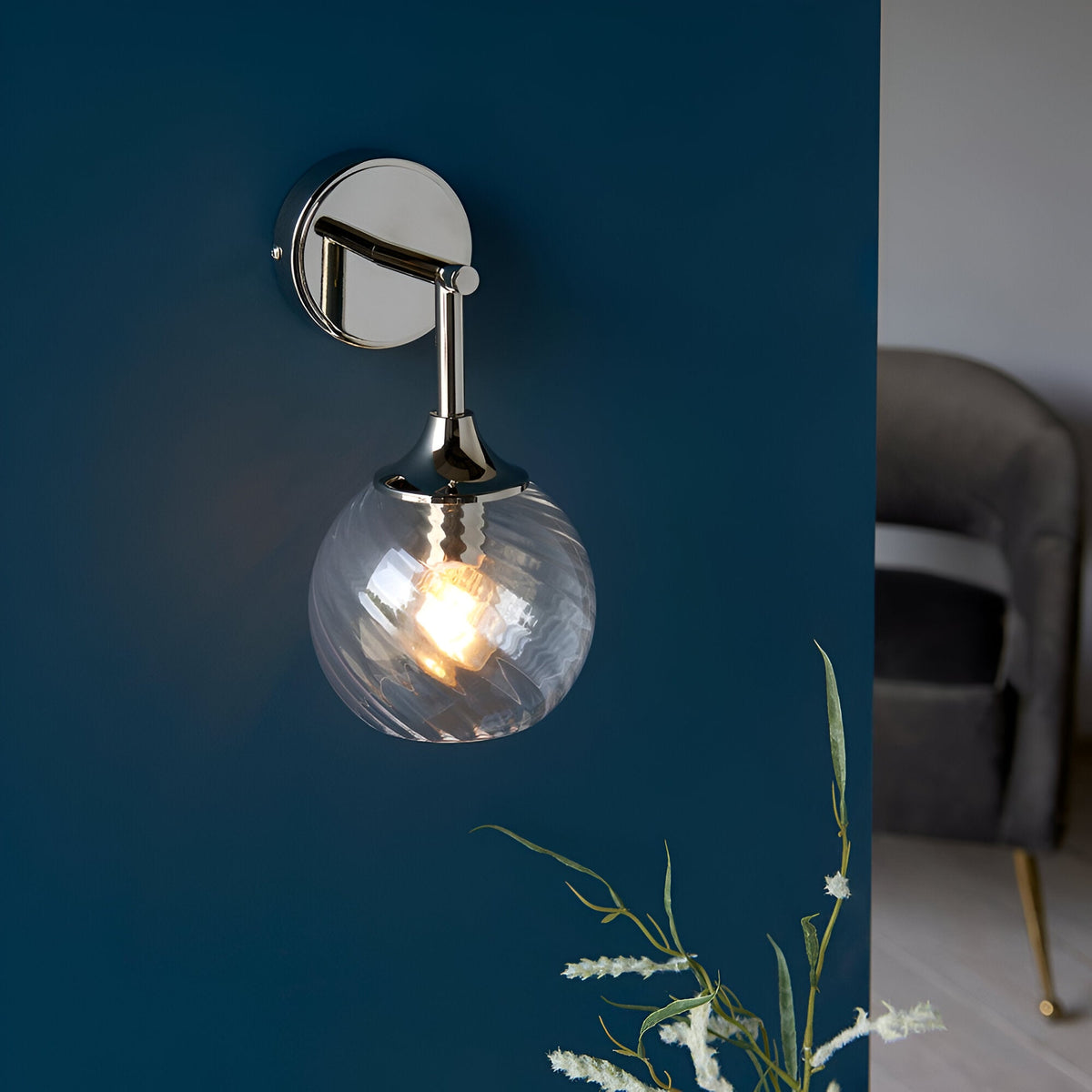 Endon Georgiana wall light with a spiral-patterned clear glass globe shade on a nickel arm, mounted on a deep blue wall in a living room setting.