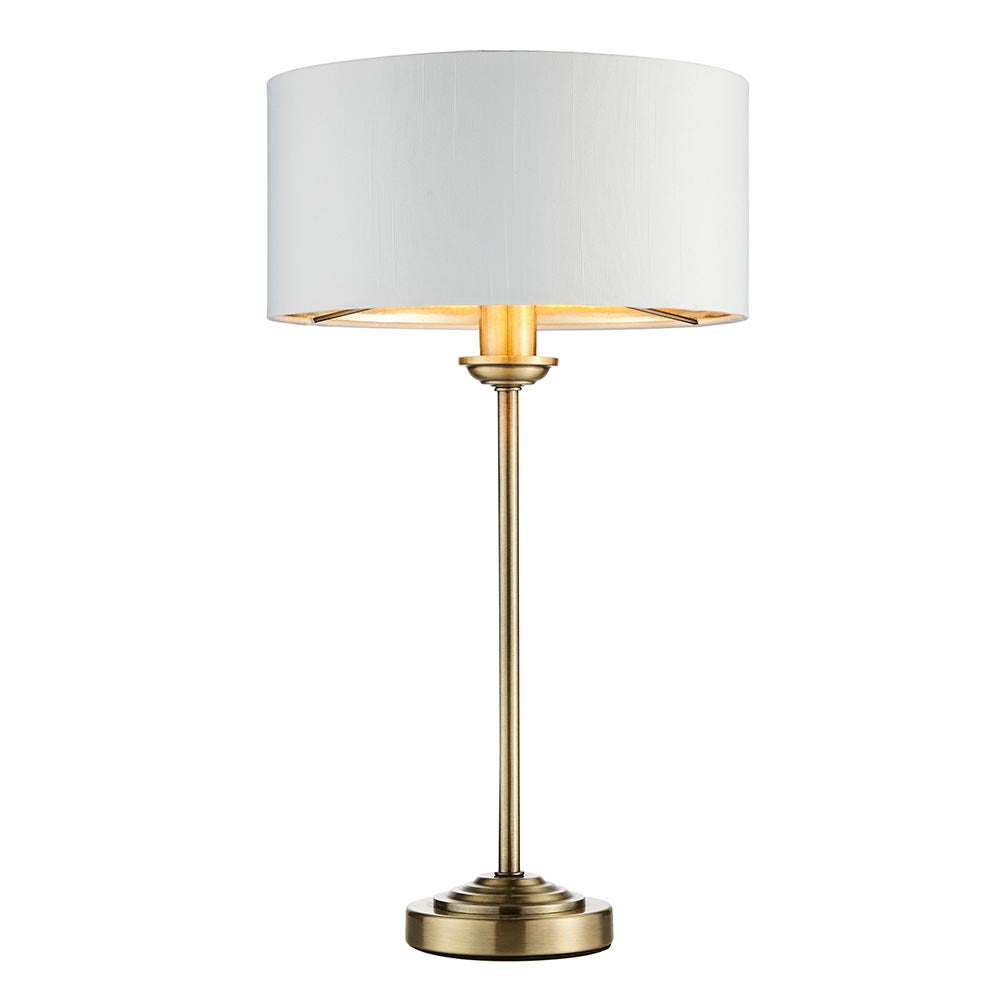 Endon Brenley table lamp with slim antique brass stem, stepped round base and white drum shade with gold metallic inner, on a plain white background.