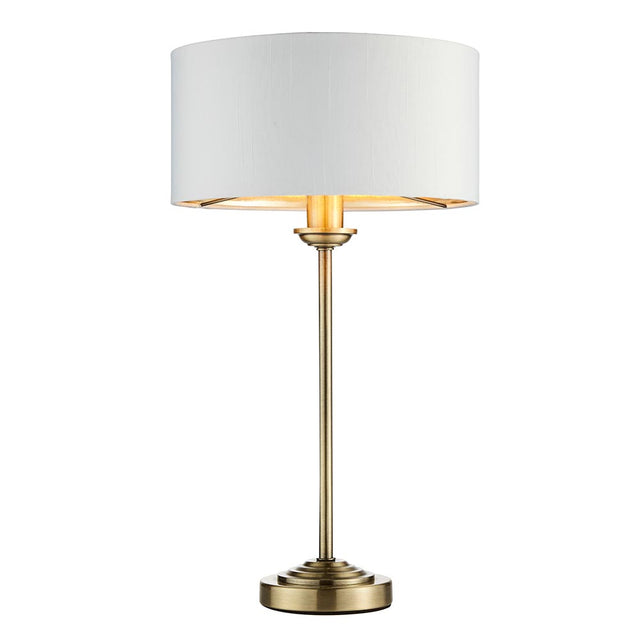 Endon Brenley table lamp with slim antique brass stem, stepped round base and white drum shade with gold metallic inner, on a plain white background.