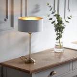 Endon Brenley table lamp with antique brass stem, white fabric shade and gold metallic inner, lit on an oak console beside a glass eucalyptus vase.
