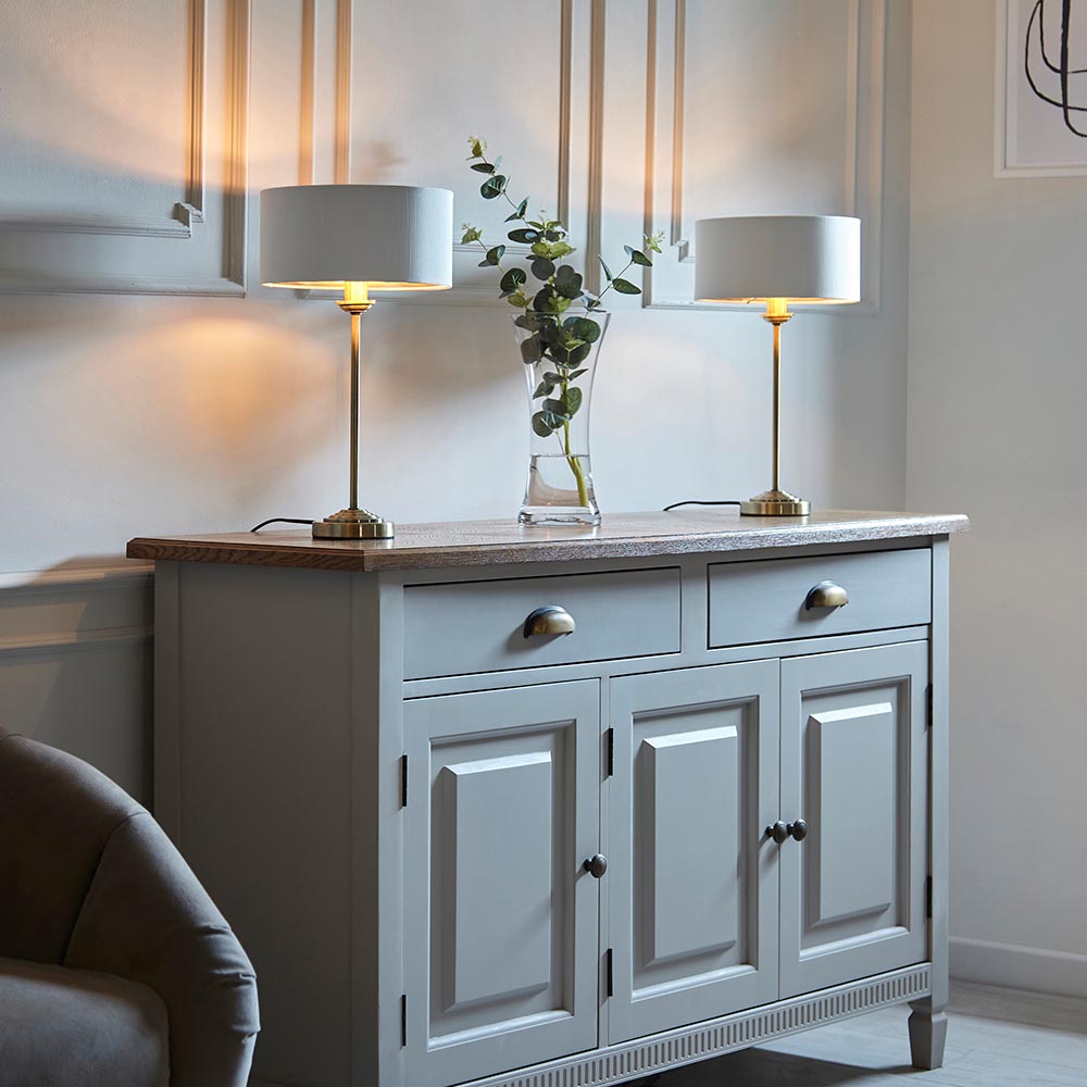 Pair of Endon Brenley table lamps with brass stems and white drum shades on a grey sideboard, beside a glass vase of eucalyptus.