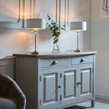 Pair of Endon Brenley table lamps with brass stems and white drum shades on a grey sideboard, beside a glass vase of eucalyptus.