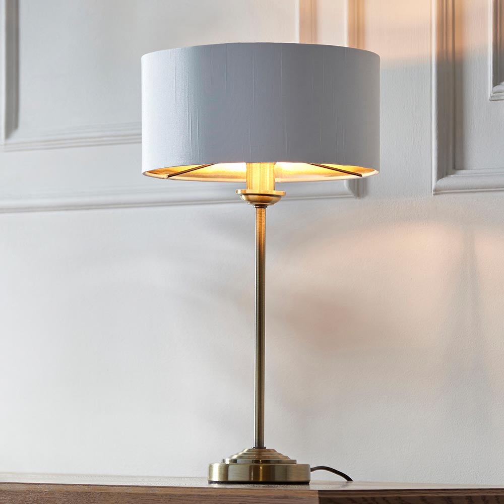 Endon Brenley table lamp with slim antique brass stem, round base and white drum shade with gold metallic inner, lit on a wood surface by panelling.
