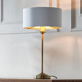 Endon Brenley table lamp with slim antique brass stem, round base and white drum shade with gold metallic inner, lit on a wood surface by panelling.