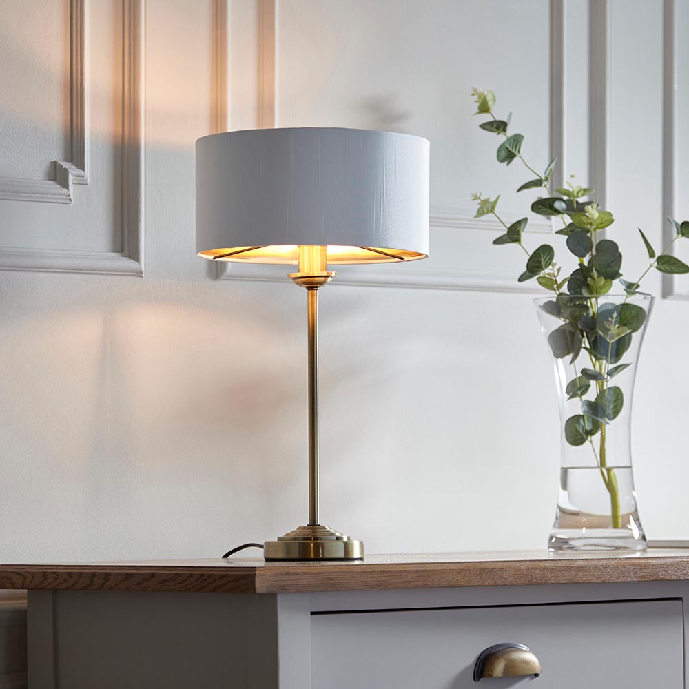 Endon Brenley table lamp with slim brass stem and white drum shade with gold inner, lit on a wood sideboard beside a glass vase of eucalyptus.
