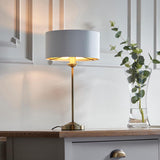 Endon Brenley table lamp with slim brass stem and white drum shade with gold inner, lit on a wood sideboard beside a glass vase of eucalyptus.