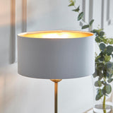 Endon Brenley table lamp lit on a brass stem with white fabric drum shade and gold metallic inner, beside eucalyptus foliage in a bright interior.