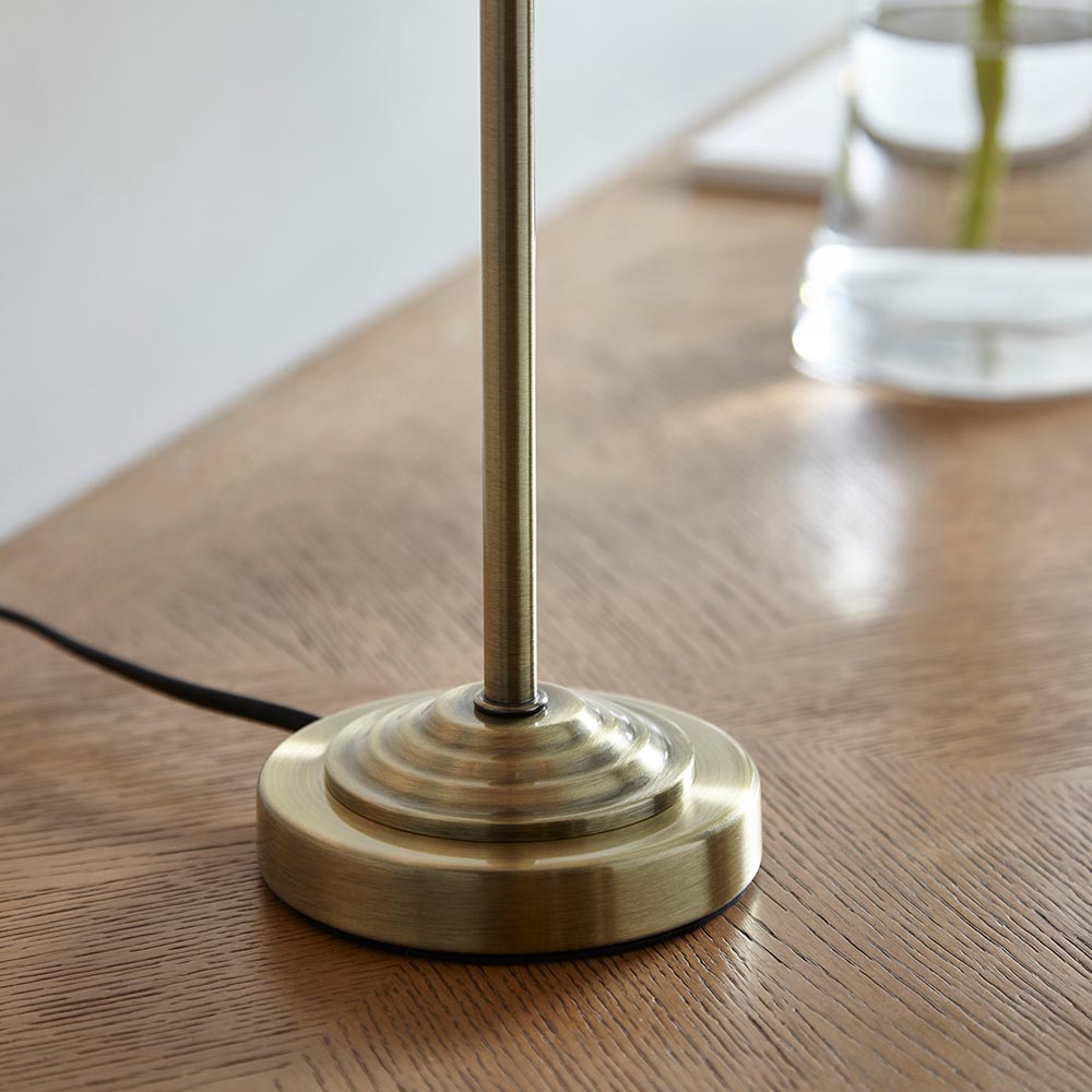 Close-up of the Endon Brenley table lamp's tiered antique brass base and slim stem on a wooden surface, with black flex trailing to the side.