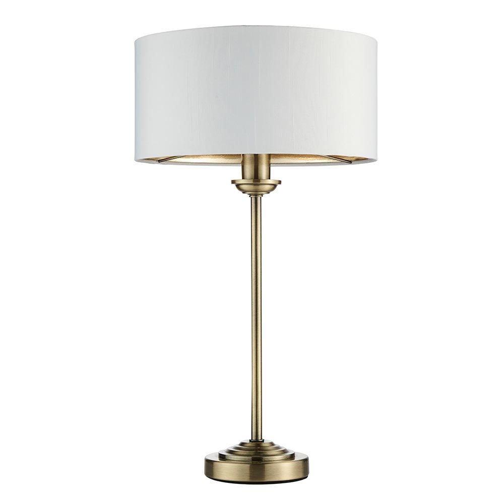 Endon Brenley table lamp with slim antique brass stem, stepped base and white drum shade with gold metallic inner, shown on a plain background.