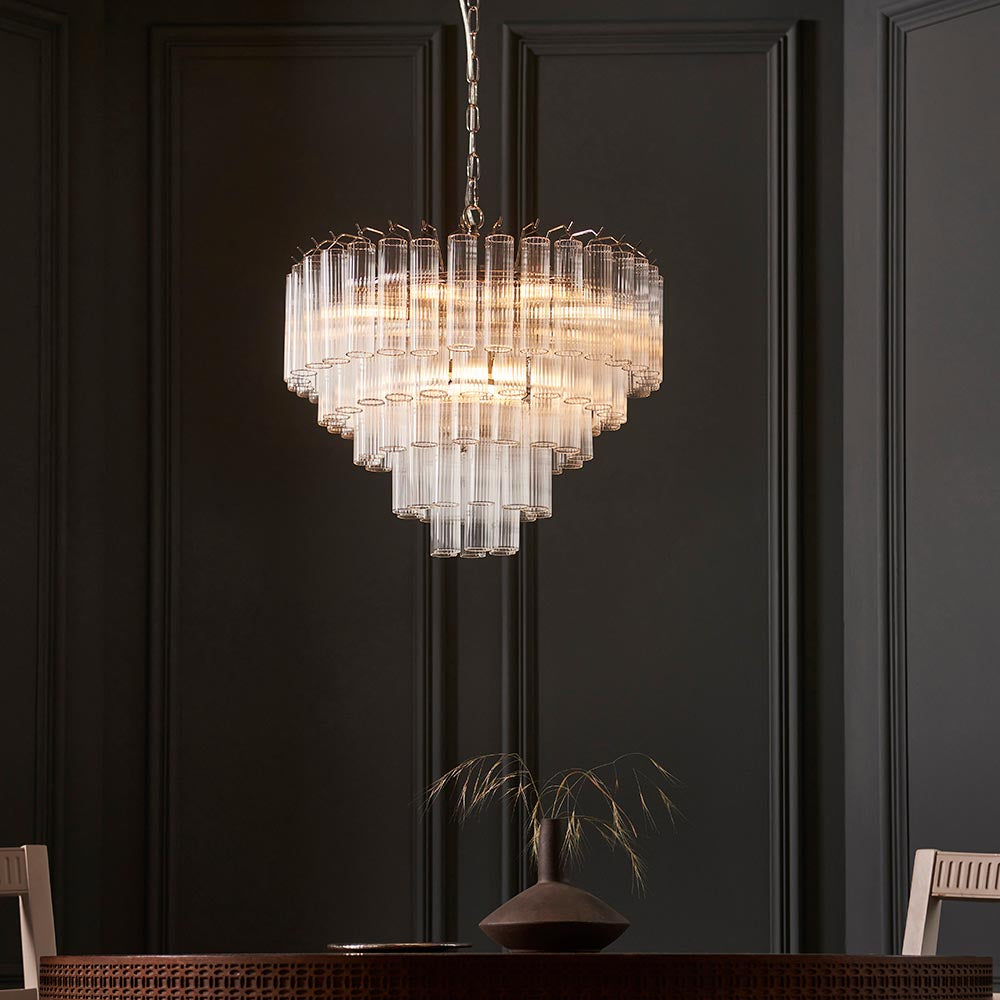 Endon Cranshaw 12 light tiered pendant in polished chrome with ribbed clear glass tubes, hanging from a chain above a dining table against a dark panelled wall.