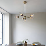 Caldworth 6 Light Pendant in brass with six curved arms and clear glass shades, hanging above a dining table in a neutral-toned room.
