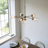Caldworth 6 Light Pendant in brass with curved arms and clear glass shades, hanging above a dining table in a light-filled room.