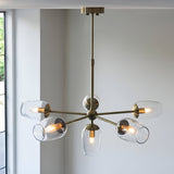 Caldworth 6 Light Pendant in brass, six curved arms with clear glass shades and exposed bulbs, hung from a telescopic stem in a light-coloured room.
