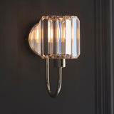 Endon Clementine wall light in nickel with a faceted clear cut glass shade and curved plated metal arm, lit against a dark grey wall.