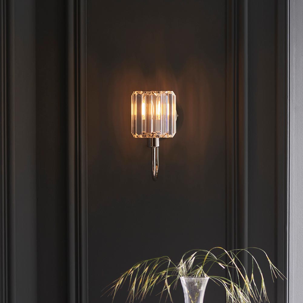 Endon Clementine wall light in nickel with a faceted clear glass shade and lit bulb, mounted on a dark panelled wall above a grass stem in a vase.