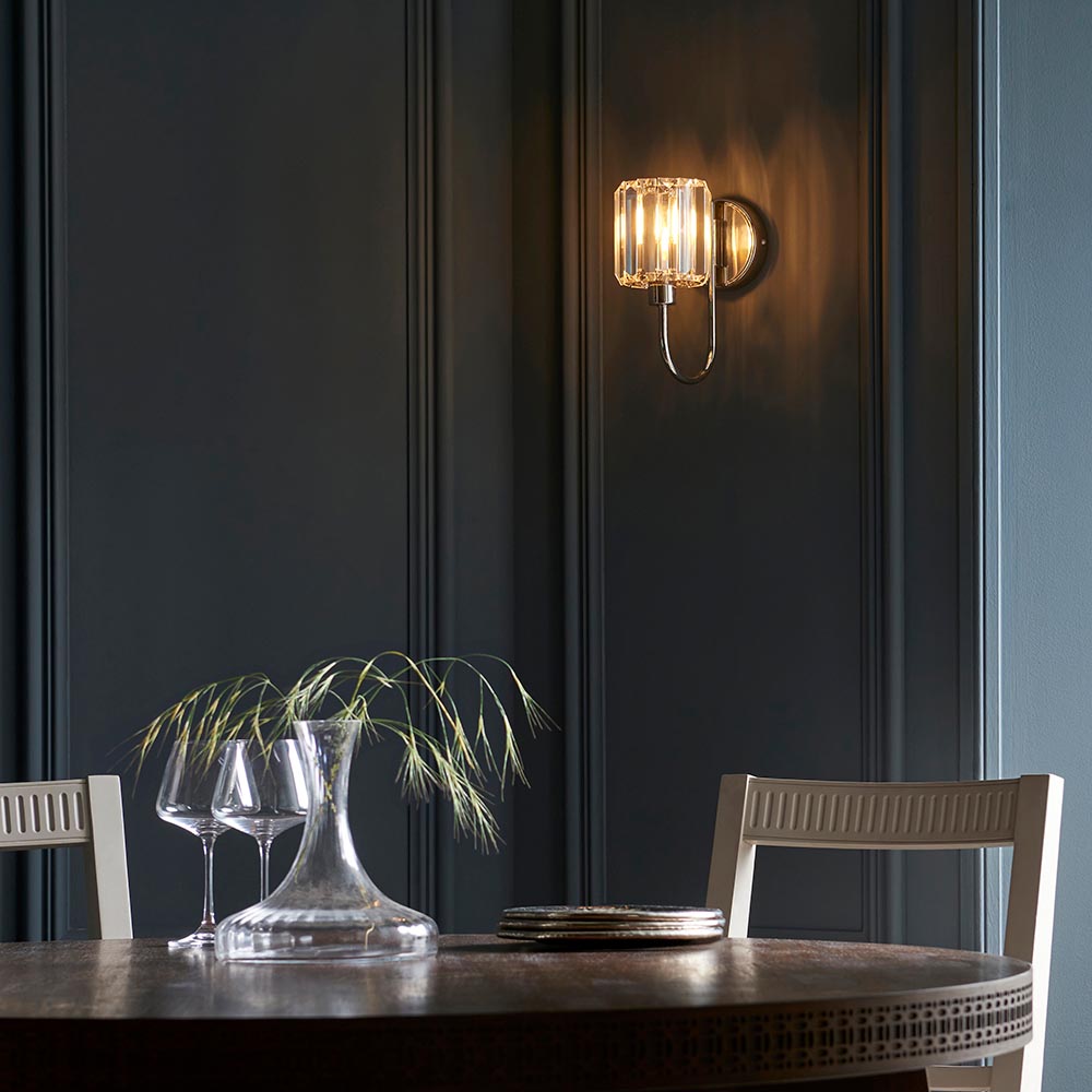 Endon Clementine wall light in nickel with a faceted clear glass shade, illuminated above a dining table set against a dark panelled wall.