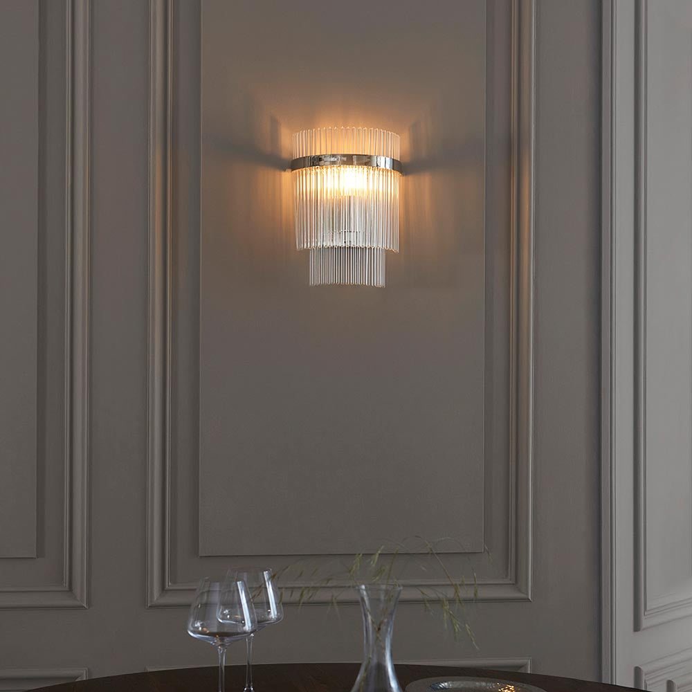 Endon Beaumont wall light in nickel with tiered clear glass rods, illuminated on a panelled grey wall above a dining table with glassware.