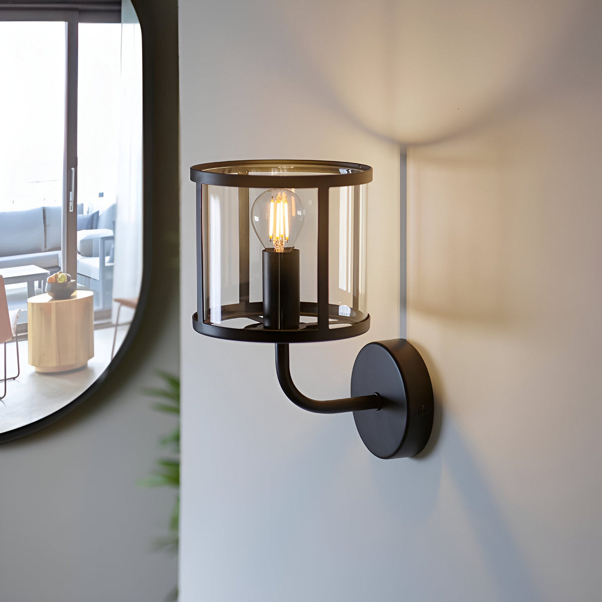 Endon Ellery wall light in matt black with a clear cylindrical glass shade and E14 filament bulb, mounted beside a round mirror in a bright interior.