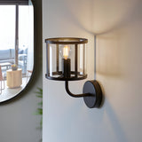 Endon Ellery wall light in matt black with a clear cylindrical glass shade and E14 filament bulb, mounted beside a round mirror in a bright interior.