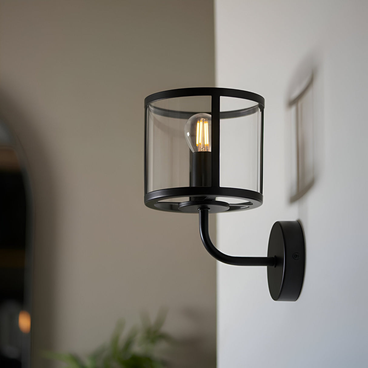 Endon Ellery wall light in matt black with a cylindrical clear glass shade and E14 filament bulb, mounted on a hallway wall beside a framed picture.