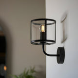 Endon Ellery wall light in matt black with a cylindrical clear glass shade and E14 filament bulb, mounted on a hallway wall beside a framed picture.