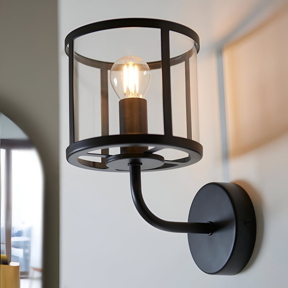 Endon Ellery wall light in matt black with a clear cylindrical glass shade and E14 filament bulb, mounted on a light interior wall.