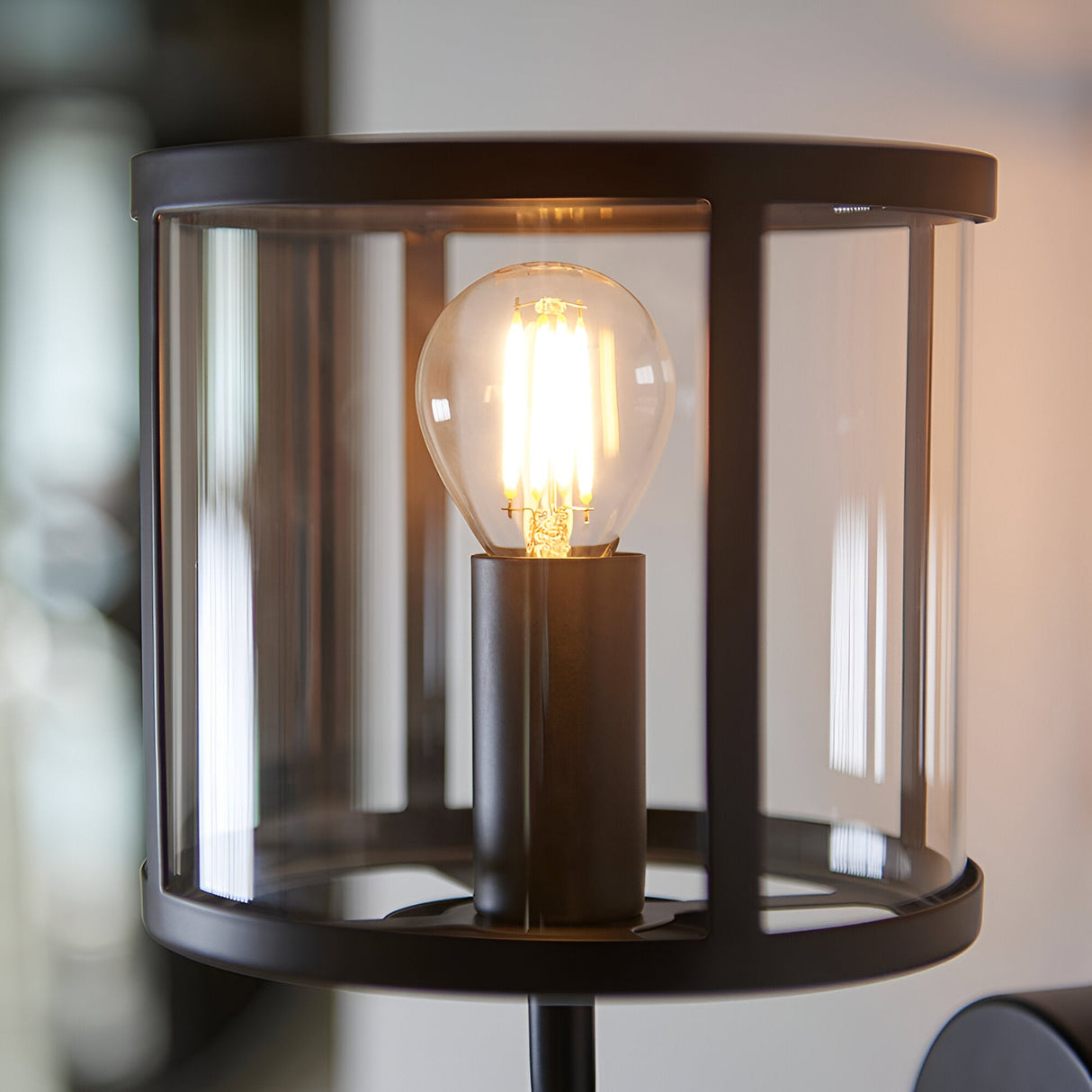 Endon Ellery wall light close-up showing the clear glass cylindrical shade, matt black metal frame and lit E14 LED filament bulb.