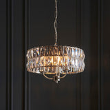 Bevmont 3 Light Pendant in nickel with a drum shade of faceted crystal glass on a chain suspension, hanging against a dark panelled wall.