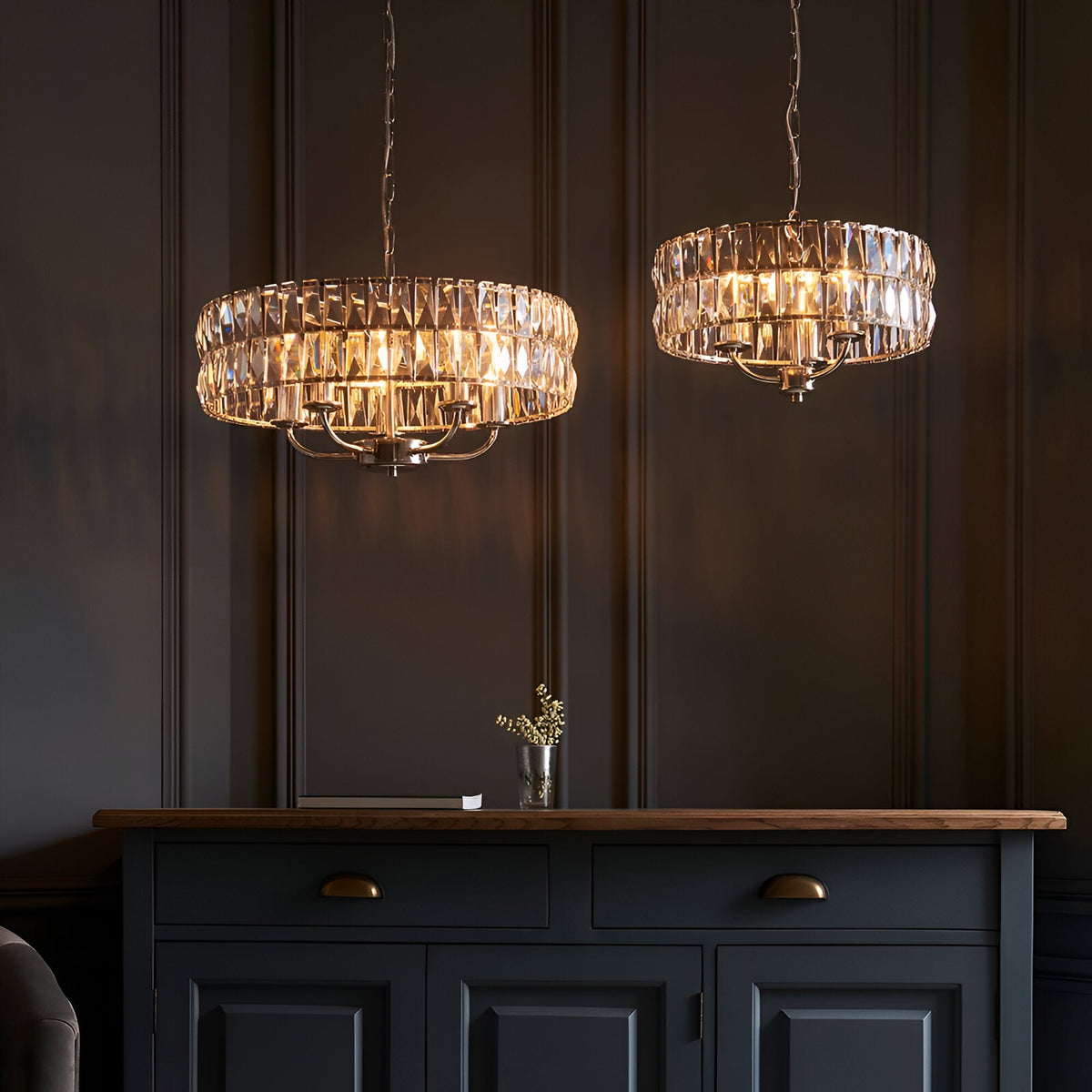 Endon Bevmont 5 light pendants in nickel with cut faceted glass drum shades, hung as a pair above a dark blue sideboard in a panelled room.