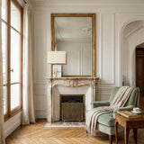 Elegant living room with a marble fireplace, ornate mirror, sage green armchair draped with a beige throw, wooden side table with tea and books, and the Veridan 1 Light Framed Marble Table Lamp - Brass & Grey by tall curtained windows and white paneled walls.