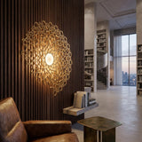 A modern living space featuring a Notredame LED Large Wall Light - Gold with intricate geometric design, a brown leather armchair, metallic side table, and a spiral staircase with built-in bookshelves.
